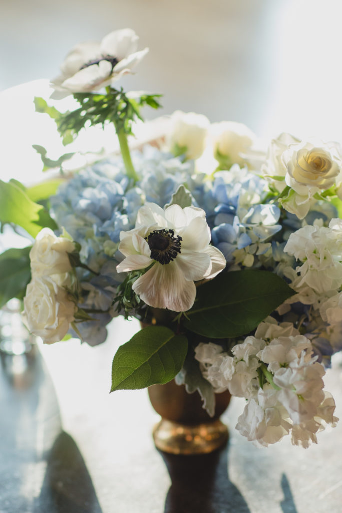 Low and soft hued arrangement in a brass vase with anemones, blue hydrangea, spray roses, stock, and dusty miller. 