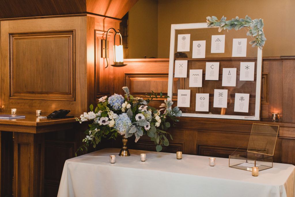Escort table with white framed glass decoration, tea light candles in mercury glass, and a garden-inspired arrangement of blue hydrangea, anemones, dusty miller, eucalyptus, and stock. 