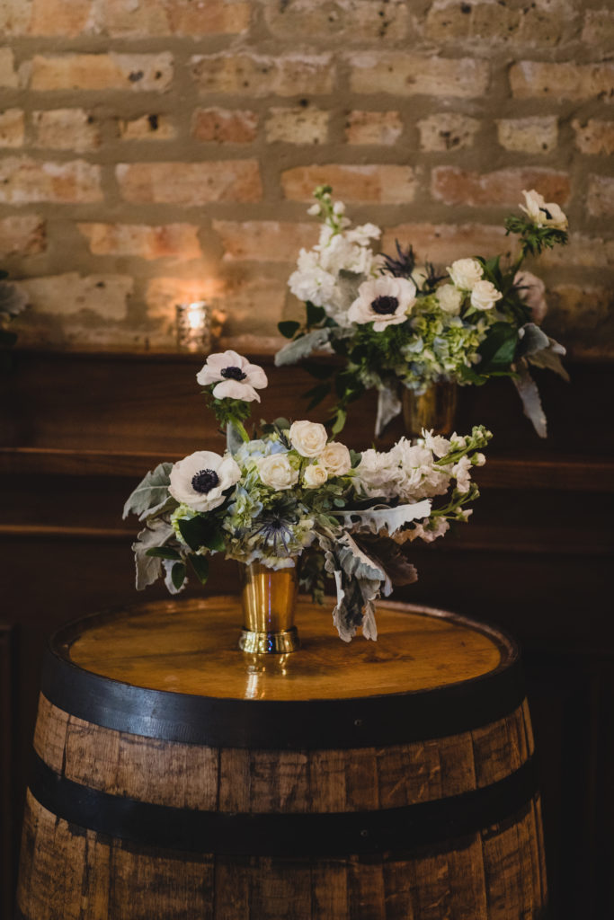 Soft hued arrangements at Revolution Brewing on a beer barrel for a cozy wedding ceremony; flowers included white anemones, blue hydrangea, spray roses, stock, dusty miller, and thistle in brass vases. 