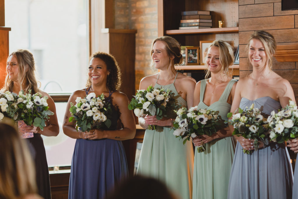 Smiling bridesmaids at Revolution Brewing Chicago, wearing a palette of sage green and dusty blues with bouquets of anemone, ranunculus, spray roses, eucalyptus, dusty miller, and thistle. 