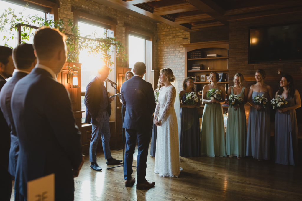 Intimate wedding ceremony with a minimal, industrial alter lined in foliage and a soft color palette of sage green, dusty blue, and ivory at Revolution Brewing in Chicago. 