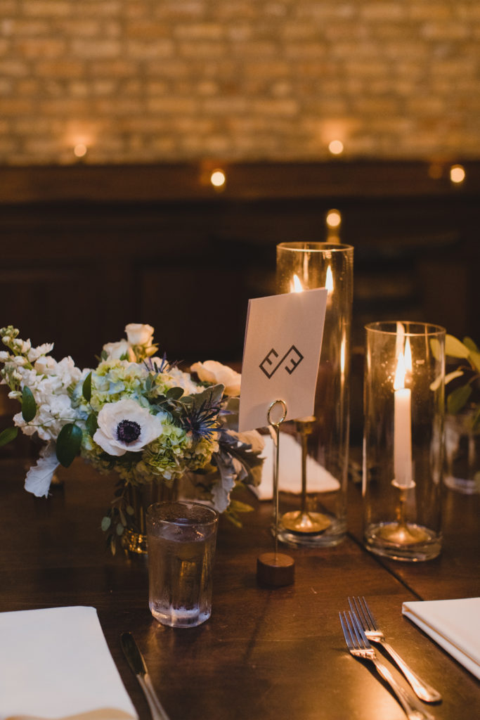 Green and blue hydrangea, thistle, white anemones, stock, and spray roses in short brass vases with taper candles in hurricane candleholders for a Revolution Brewing wedding ceremony and reception. 