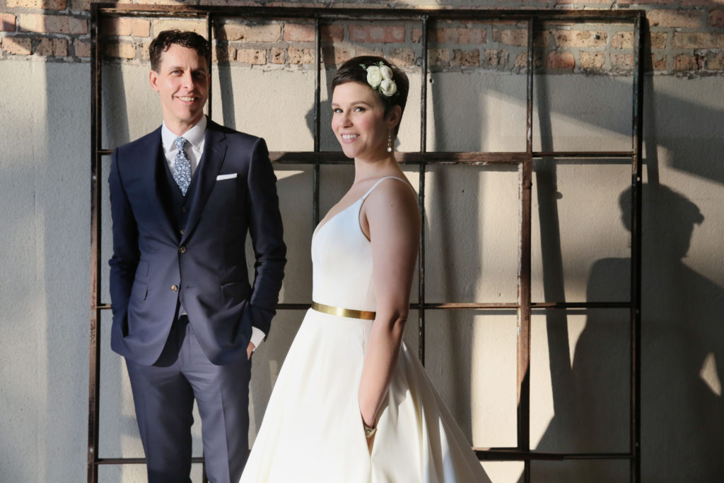 Bride and groom photoshoot with industrial background; bride wears white ranunculus flower clip with a pixie cut and golden belt.