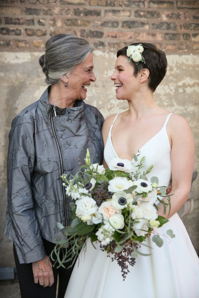 Smiling bride and mother-in-law portrait with a lush bouquet of garden roses, anemone, lisianthus, eucalyptus, and berries in monochromatic tones of white and sage green; bride wore a white ranunculus flower clip with her pixie cut. Wedding day at Artifact Events in Chicago.