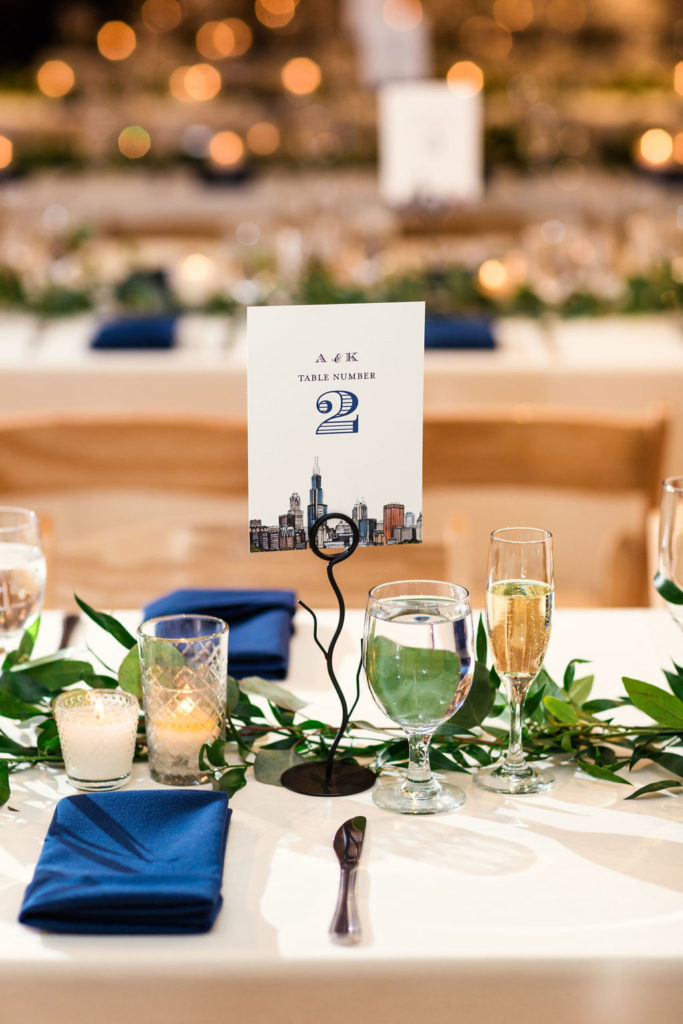 Table number cards with Chicago skyline for this all-greenery winter wedding; reception included blue and white table linens and foliage garland.