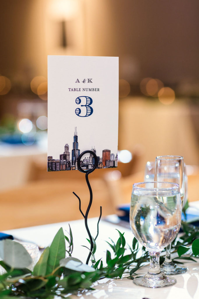 Table number cards with Chicago skyline for this all-greenery winter wedding; reception included blue and white table linens and foliage garland.