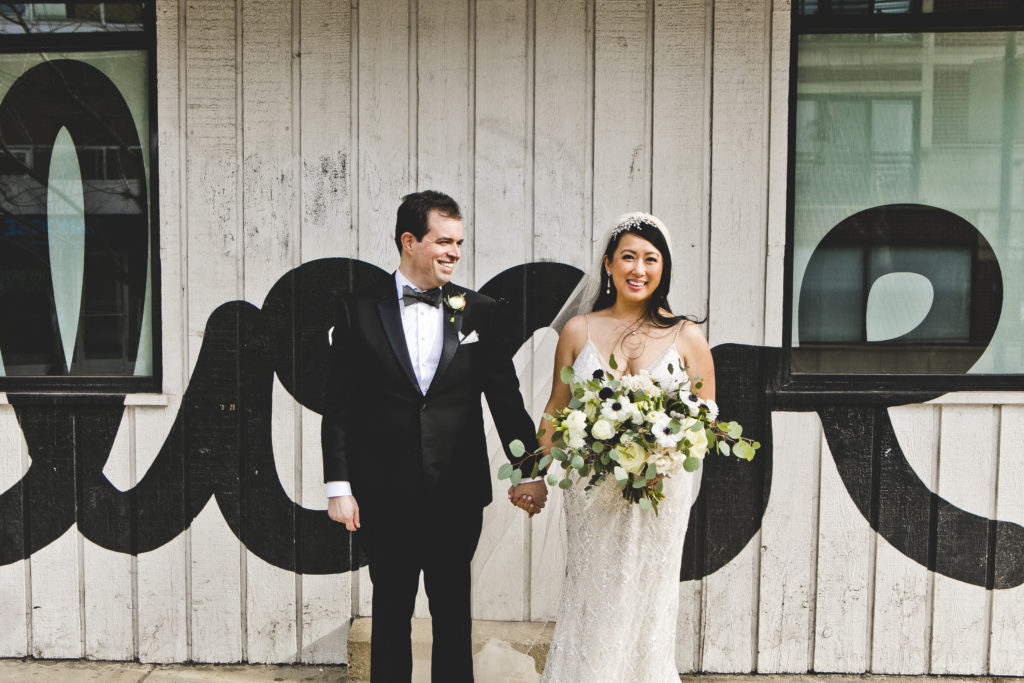 Bride and groom in a beaded grown and classic tux standing in front of 'love' street art, holding a monochromatic, loose gardeny bouquet of ivory garden roses, anemones, eucalyptus, and plum scabiosa. 