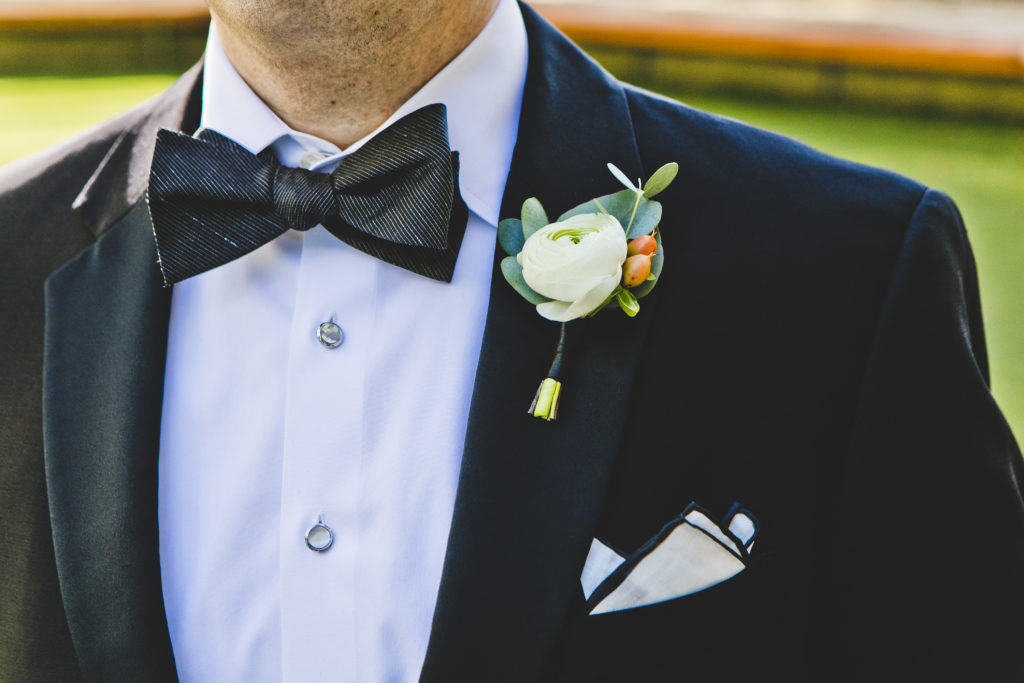 Groom with a classic bowtie wearing a classic ivory ranunculus boutonniere with peach hypericum berries and eucalyptus for a sunny wedding at Greenhouse Loft.