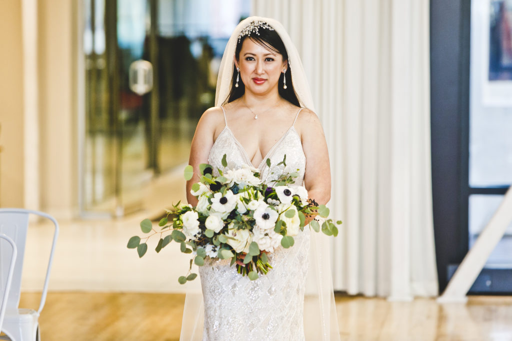 Bride walking down the aisle holding a loose and gardeny monochromatic bouquet of anemones garden rose, and eucalyptus bouquet.