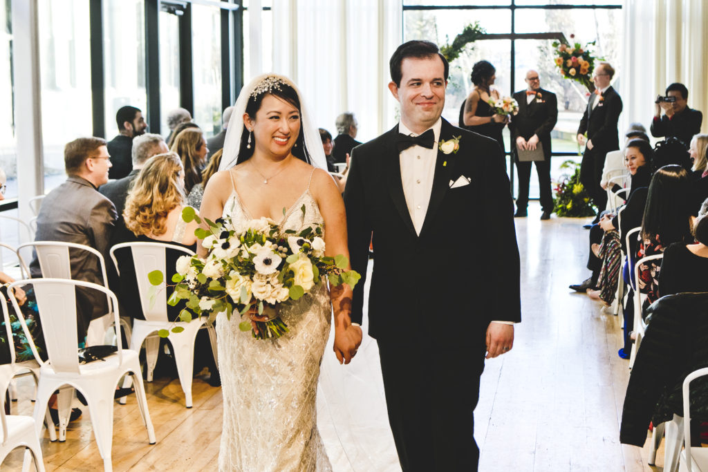 Bride and groom during wedding ceremony at Greenhouse Loft in front of contemporary, geometric alter with wildflowers, orange garden roses, ranunculus, and protea. 