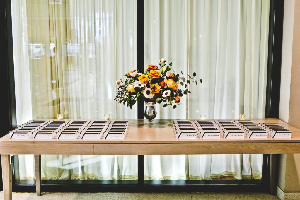 Contemporary escort card table with acrylic cards at Greenhouse Loft with a vivid centerpiece of white anemones, orange garden roses, spray roses and foliage in a vintage-style vase. 