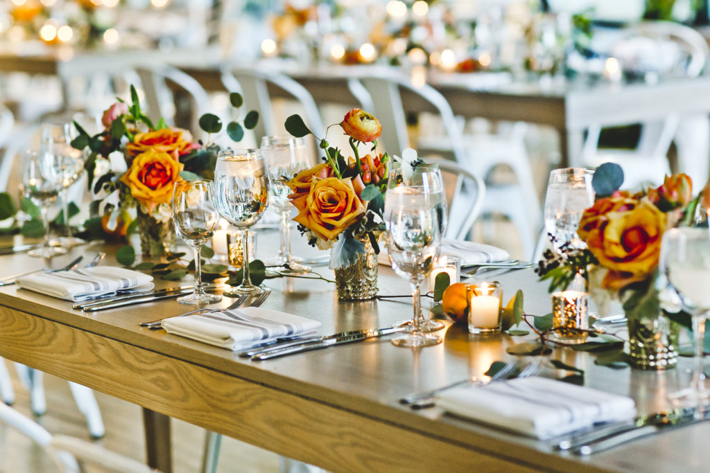 Sun-filled Greenhouse Loft wedding reception with elegant details of orange garden roses and ranunculus in mercury glass vases lining the tables with industrial farmhouse chairs.