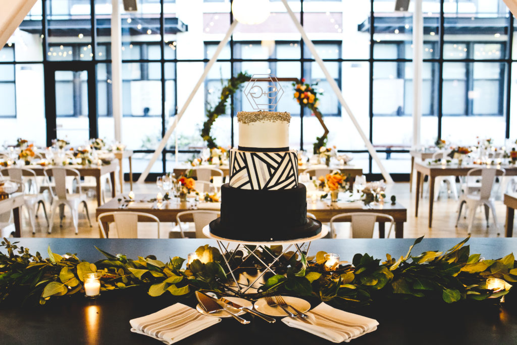 The couple's stunning graphic black and white three-tiered cake with monogrammed topper at Greenhouse Loft Chicago, on display with votive candles and lush foliage.