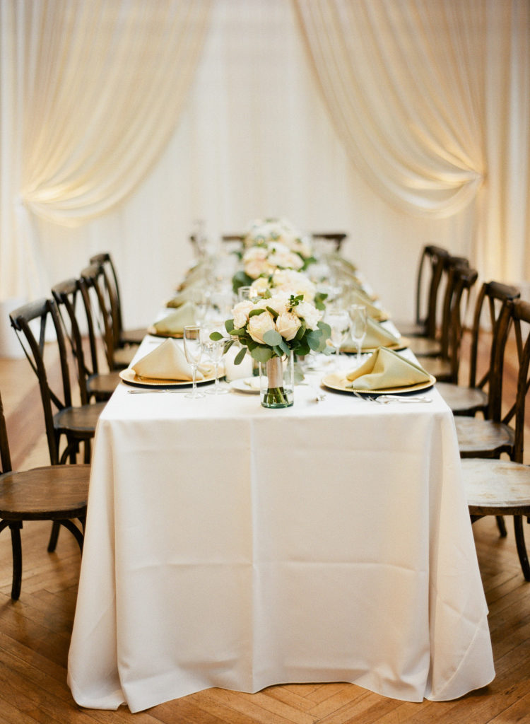 Wedding head table with white bouquets, gold plates and napkins, white table cloth, and wooden chairs at Bridgeport Art Center in Chicago.