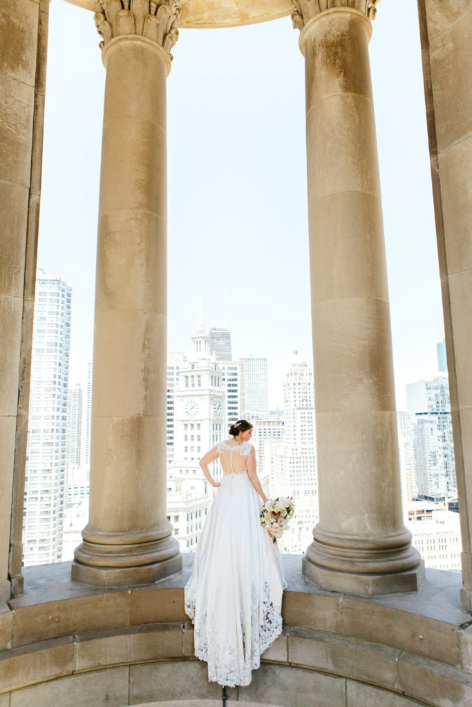 Bride posing on ledge in the city with a draping lace gown and bouquet of blush garden roses, ranuculus, and ivory peonies. 
