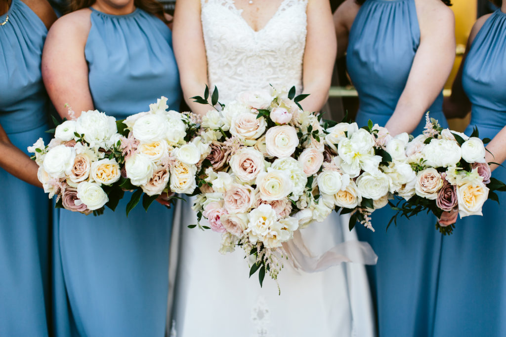 Bridesmaids in sky blue dresses with bride holding bouquets for a summer wedding; flowers include mauve and blush garden roses, peonies in pink and ivory, ranunculus, and astilbe.