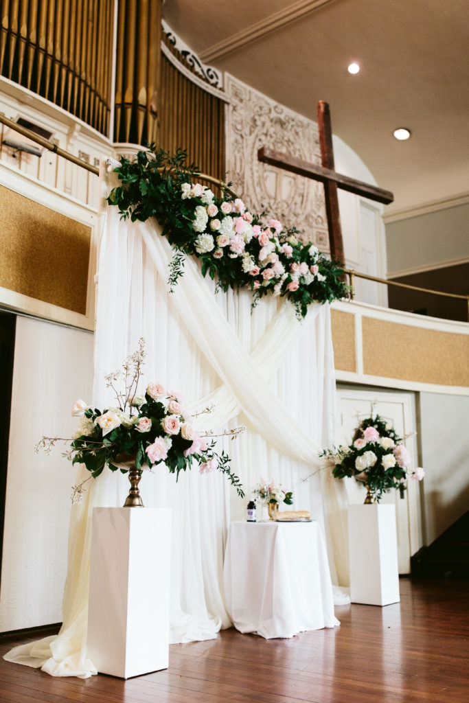 Opulent garden-style floral installation over a ceremonial backdrop for a spring wedding at Missio dei Wrigleyville in Chicago.  Flowers include hydrangea, pink peonies, and garden roses; Altar arrangements on pedestals with flowering branches add interest. 