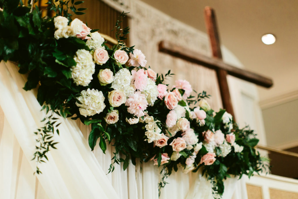 Opulent garden-style floral installation over a ceremonial backdrop for a spring wedding; flowers include hydrangea, pink peonies, and garden roses.