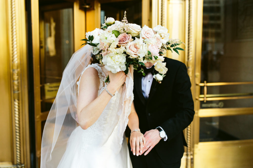 Bride and groom kissing behind a classic bouquet of ivory peonies, blush ranunculus, garden roses, and sweet pea.