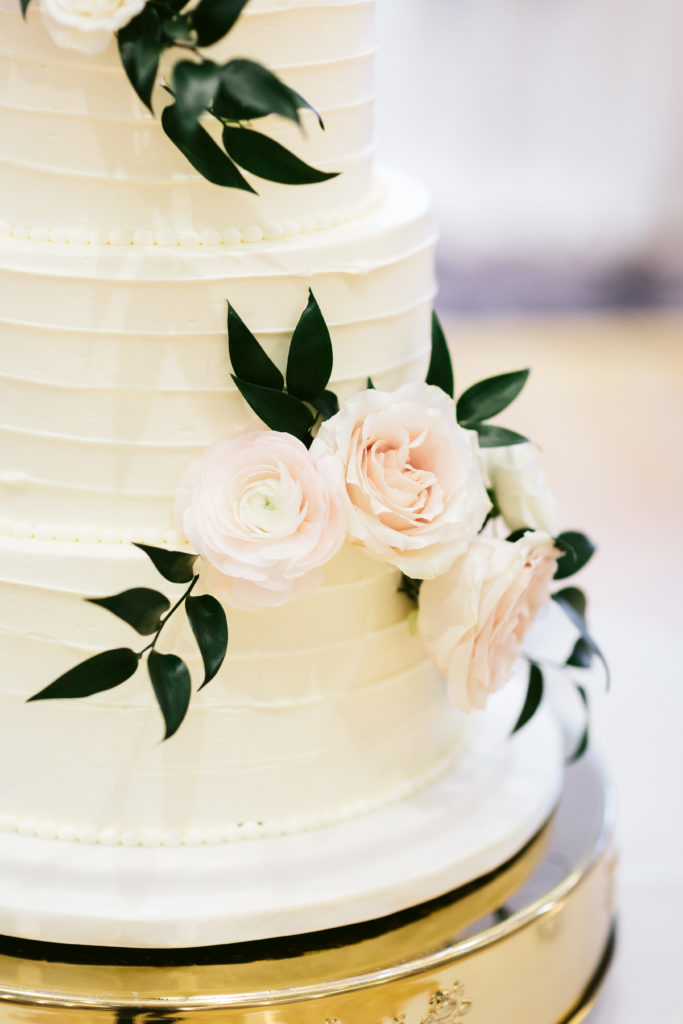 Detail of a four-tiered ivory cake with pale blush garden roses and ranunculus with foliage on a golden base. 