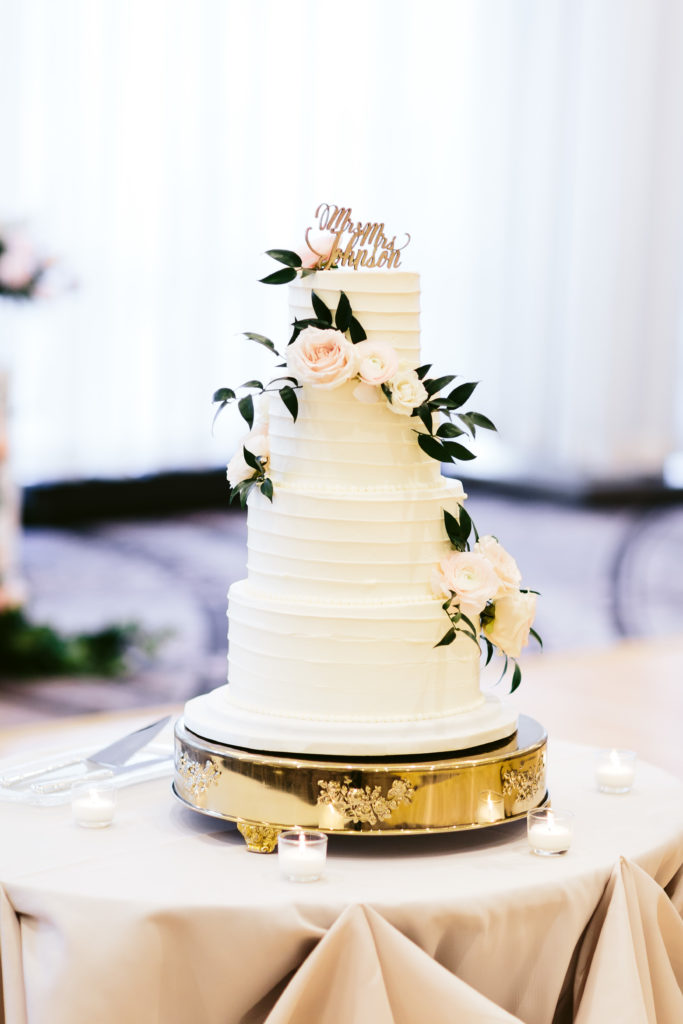 Four-tiered ivory cake with pale blush garden roses and ranunculus with foliage on a golden base. 