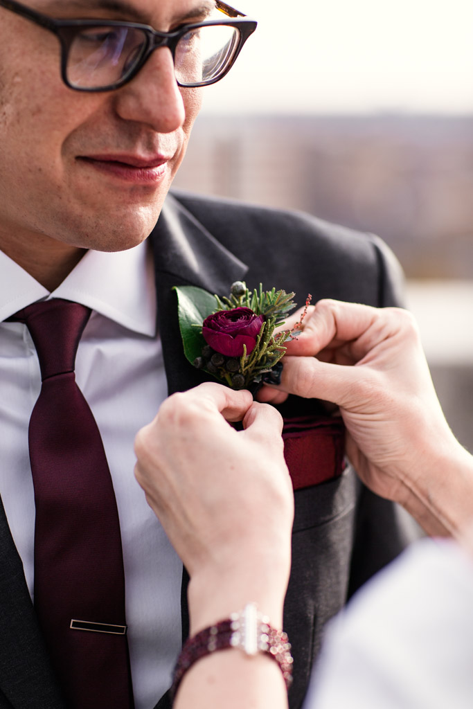 Groom gets his boutonnière with red ranunculus pinned on before the wedding ceremony. He wear a narrows dark red tie with a matching red pocket square.