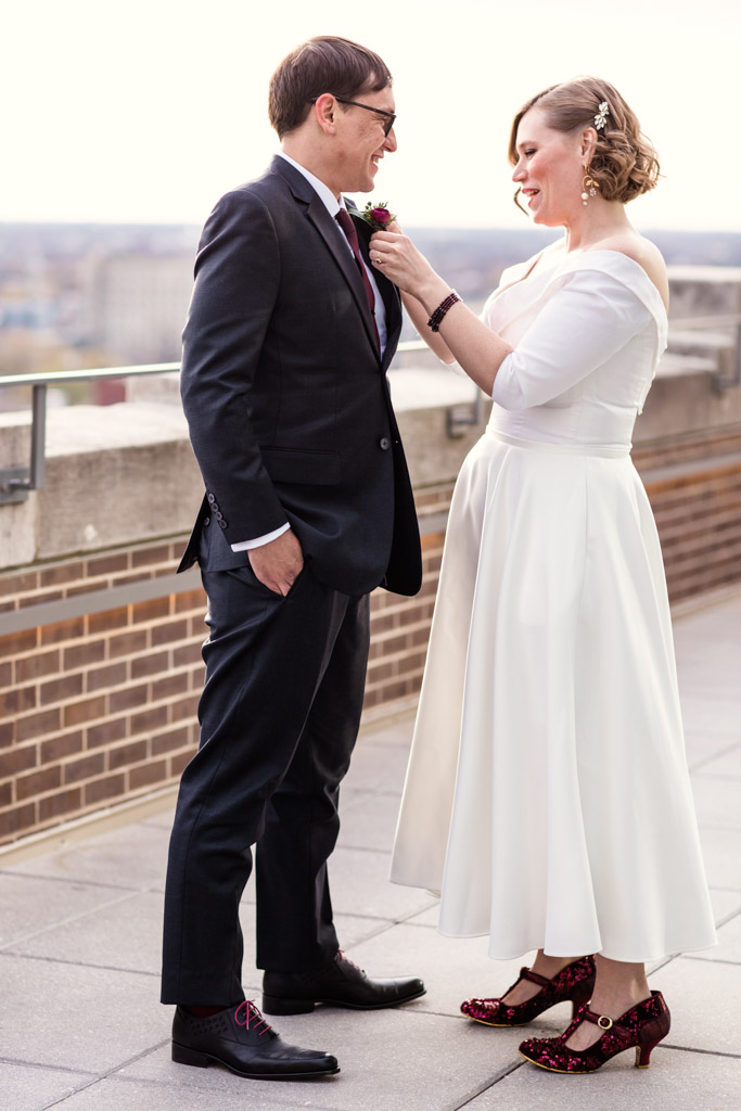 Bride and groom smile at each other as bride pins boutonnière with red ranunculus on groom. Bride wears a vintage-style, off-the-shoulder wedding dress with glitter red shoes.