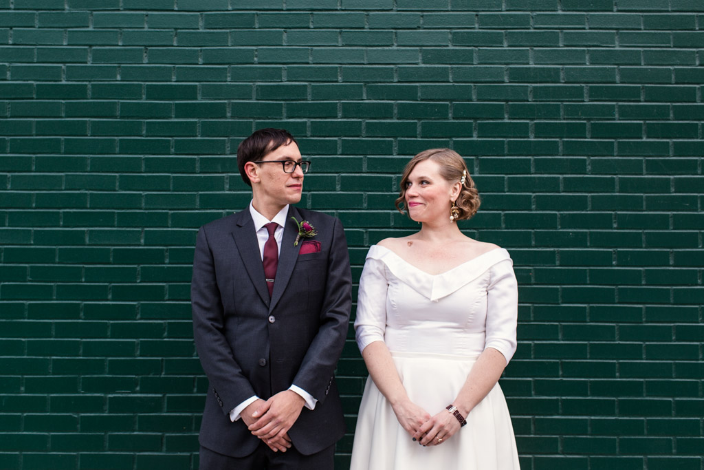 Bride and groom smile at each other while standing in front of an emerald green brick wall. Groom wears a charcoal suit with a narrow maroon tie, matching pocket square and dark red ranunculus boutonnière. Bride wears a vintage-style wedding dress with her hair pinned back.