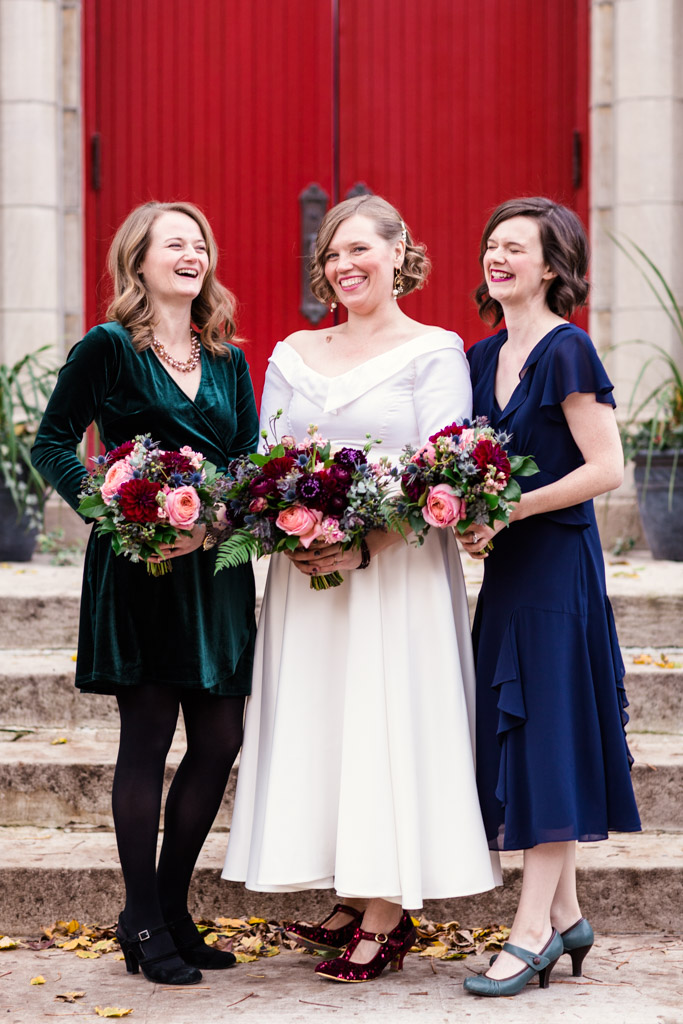 Bridal party smiles in front of red church doors for a fall ceremony featuring bouquets of pink garden roses, blue thistle, plum scabiosa, eucalyptus, fern, maroon dahlias, and berries.