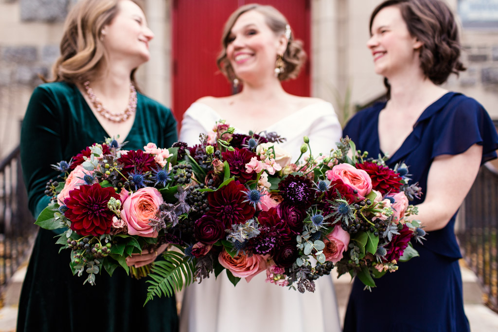 Bridal party smiles in front of red church doors for a fall ceremony featuring bouquets of pink garden roses, blue thistle, plum scabiosa, eucalyptus, fern, maroon dahlias, and berries.