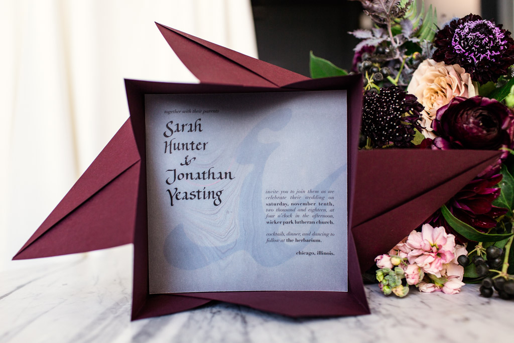 Wedding invitation in a maroon red origami envelope with the bride's bouquet of garden roses, plum scabiosa, stock, berriers, and fern behind it.