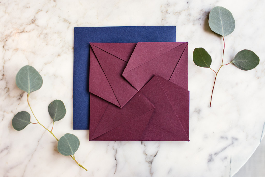 Dusty red origami envelope with a wedding invitation inside. Placed on top of a blue envelope with two eucalyptus leaves next to it and a marble background.