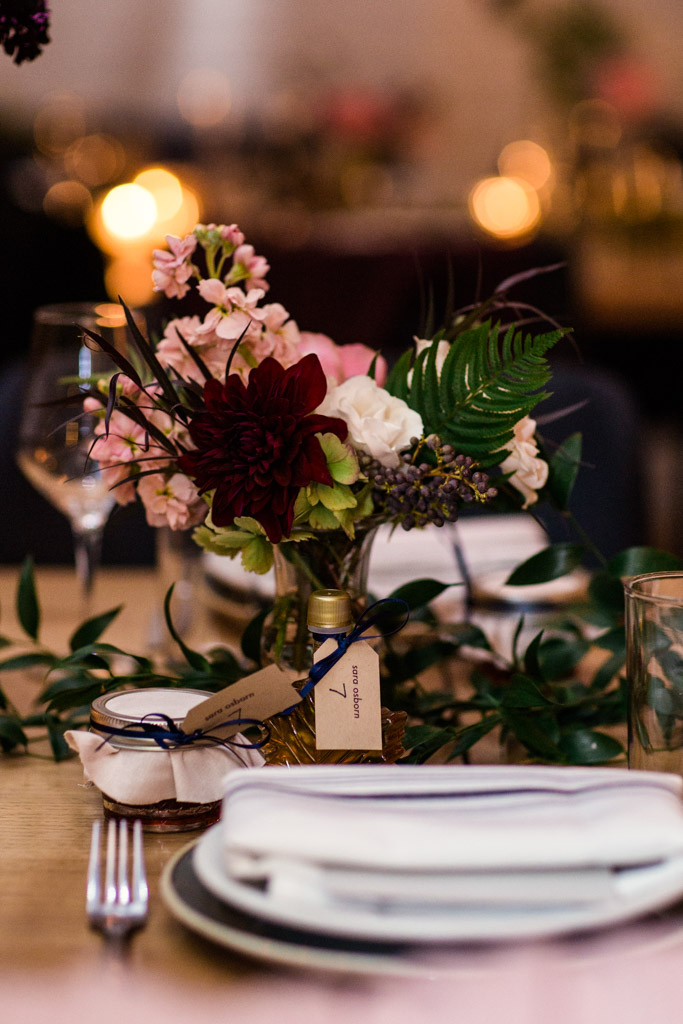 Table arrangement of maroon dahlias, fern, pink stock, and ivory spray roses placed among sprawling foliage and hand-bottled maple syrup.