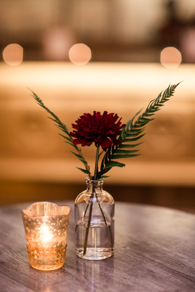 Single maroon dahlia with two fern leaves sits in a vintage-inspired glass bottle vase next to a mercury glass candle. 