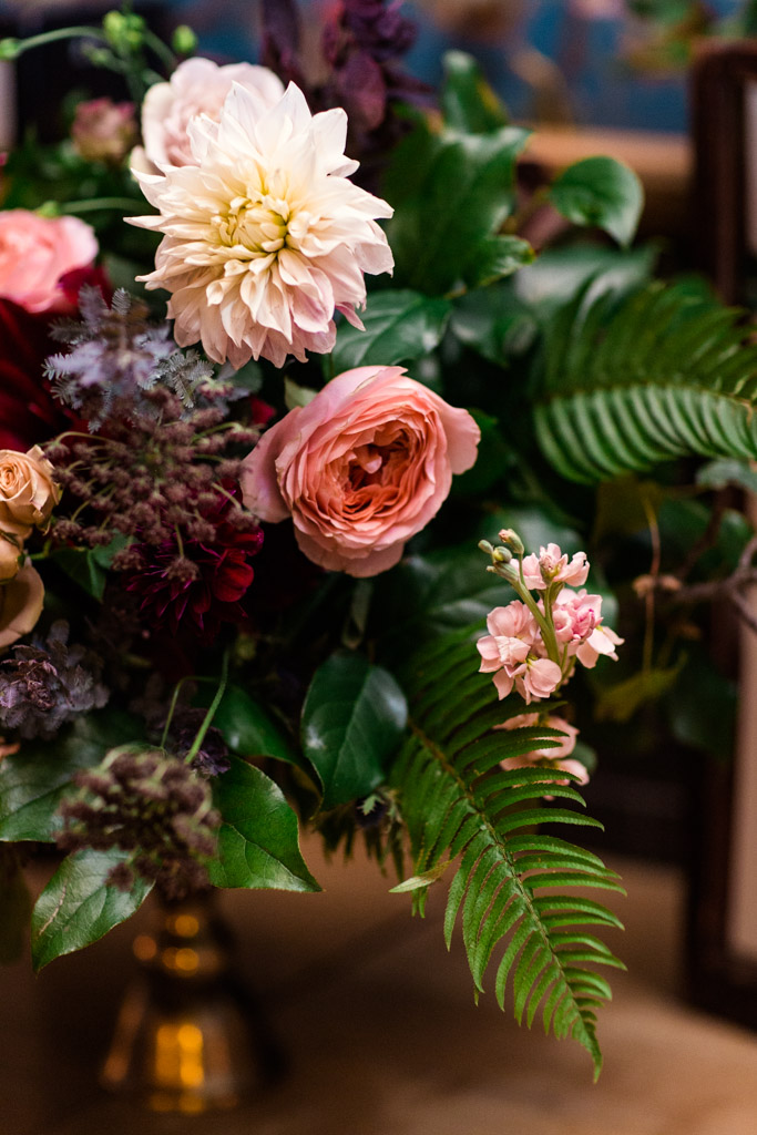Welcome table bouquet of maroon and blush dahlias, pink garden roses, black Queen Anne's lace, pink stock, pale orange spray roses, fern, and sprawling foliage. It is placed in front of decadent floral wallpaper and butterfly dioramas. 