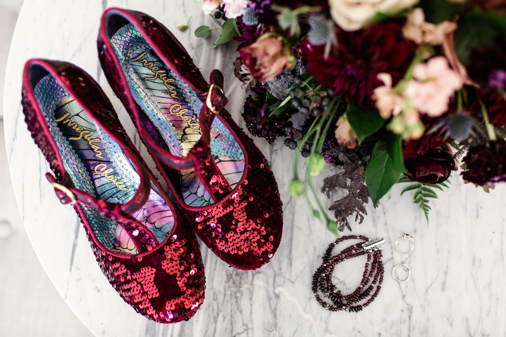 Ruby red sequin slippers for the bride's unique look on her wedding day, photographed next to her jewelry, rings, and lush bouquet of maroon dalias, spray roses, and plum scabiosa.