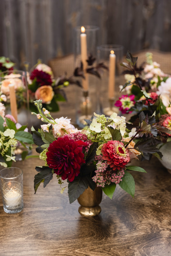 Warm and autumn inspired arrangements on the table of this rustic backyard wedding included zinnias, peach garden roses, burgundy dahlias, berries, and stock. Each was displayed within vintage inspired brass vases. 