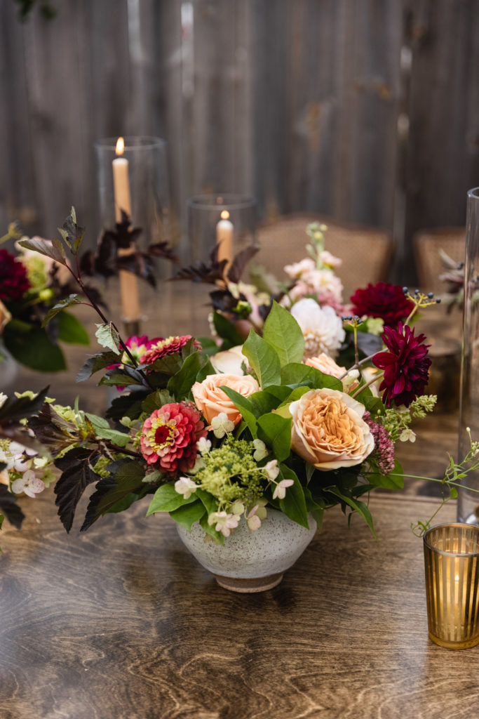Warm and autumn inspired arrangements on the table of this rustic backyard wedding included zinnias, peach garden roses, burgundy dahlias, berries, and stock. 