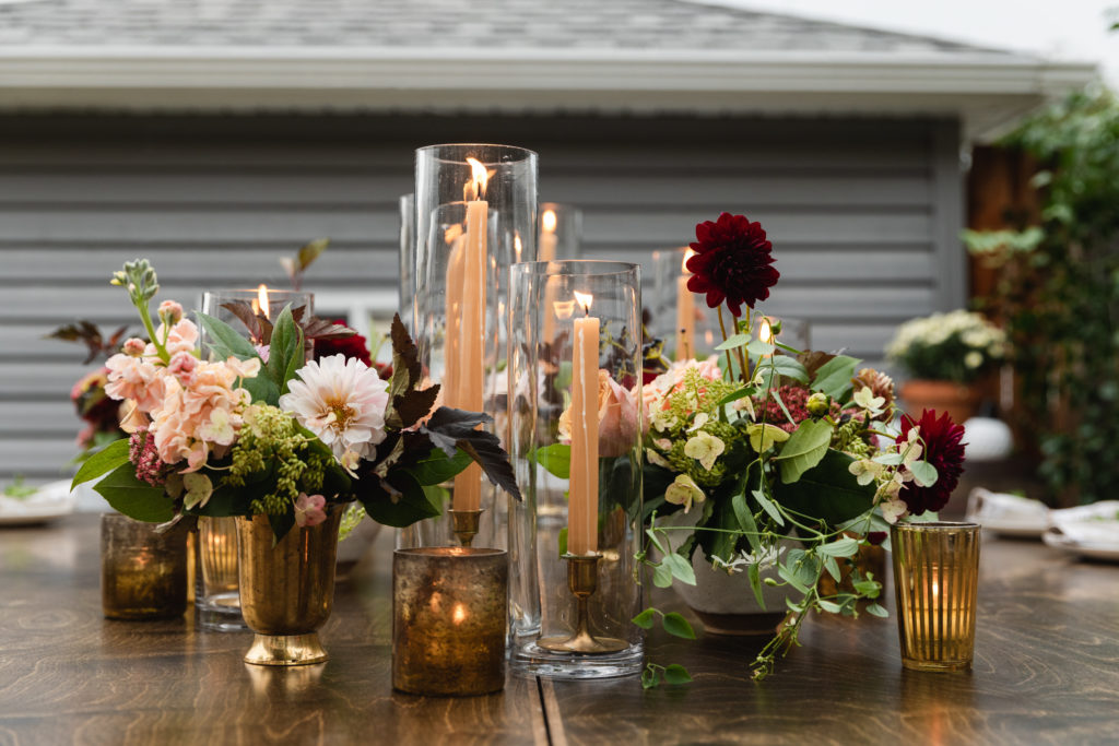 Warm and autumn inspired arrangements on the table of this rustic backyard wedding included zinnias, peach garden roses, burgundy dahlias, berries, and stock. Here they are in brass vases with honey colored taper candles and votives.