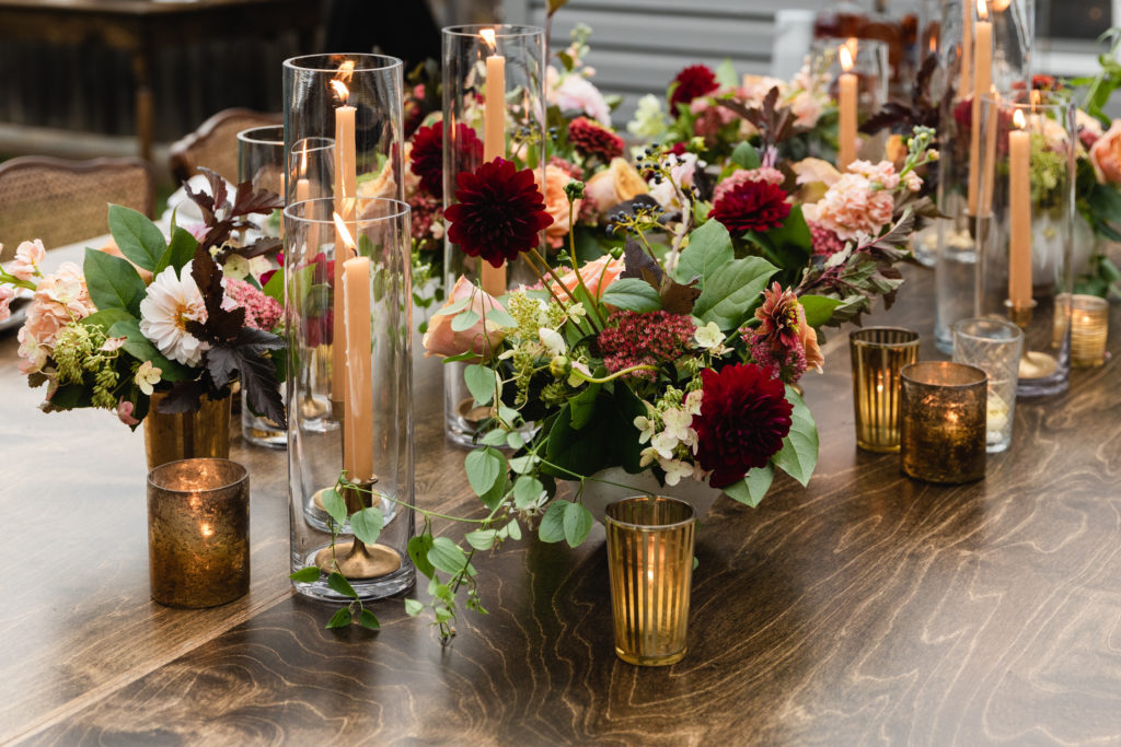 Warm and autumn inspired arrangements on the table of this rustic backyard wedding included zinnias, peach garden roses, burgundy dahlias, berries, and stock. Here they are in brass vases with honey colored taper candles and votives.