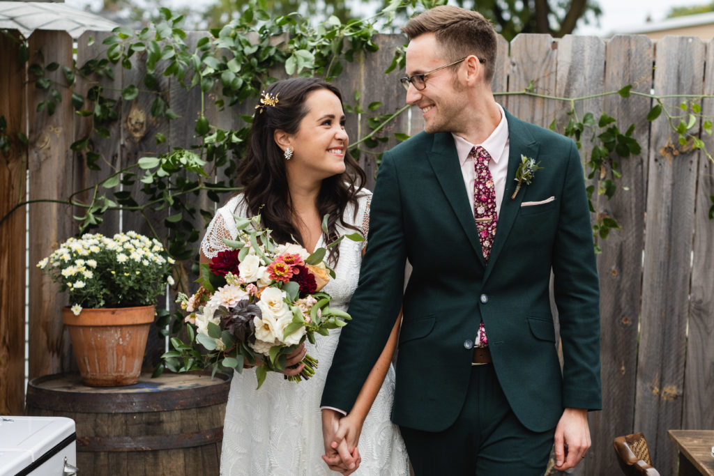 Smiling bride and groom at a micro backyard early fall wedding. Bride's bouquet features garden roses, blush and burgundy dahlias, pink zinnias, ivory majolica roses, eucalyptus, and stock.