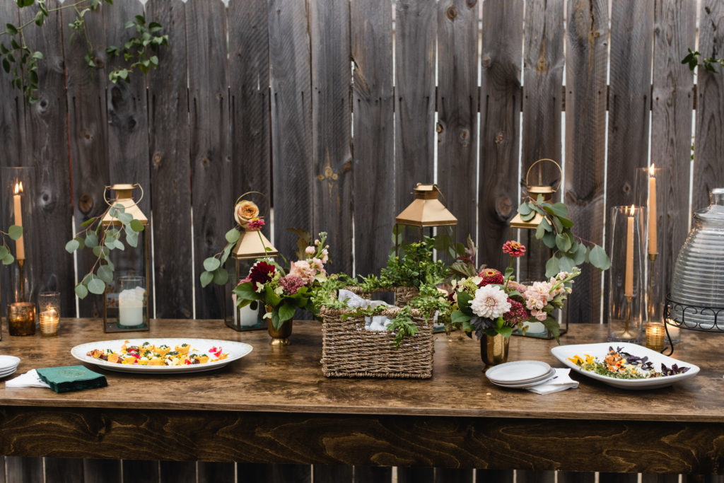Warm and autumn inspired arrangements on the table of this rustic backyard wedding included zinnias, peach garden roses, burgundy dahlias, berries, and stock. Here they are in brass vases with honey colored taper candles and lanterns adorned with eucalyptus branches.
