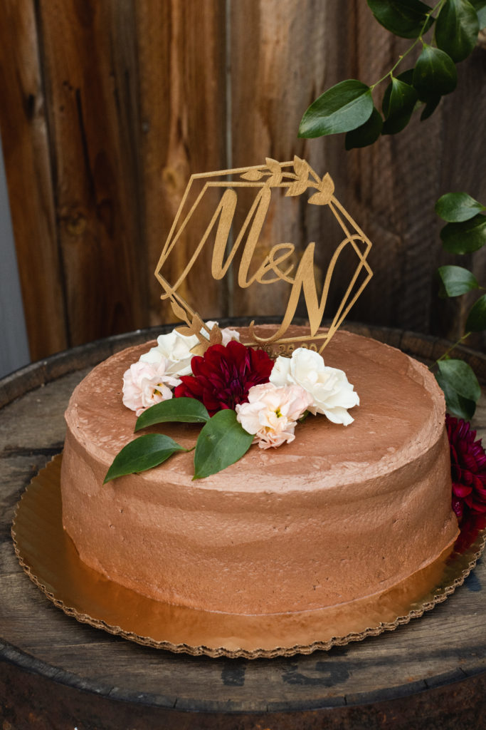 A simple chocolate frosted cake adorned with majolica roses, a burgundy dahlia, and a geometric topper with calligraphy initials.
