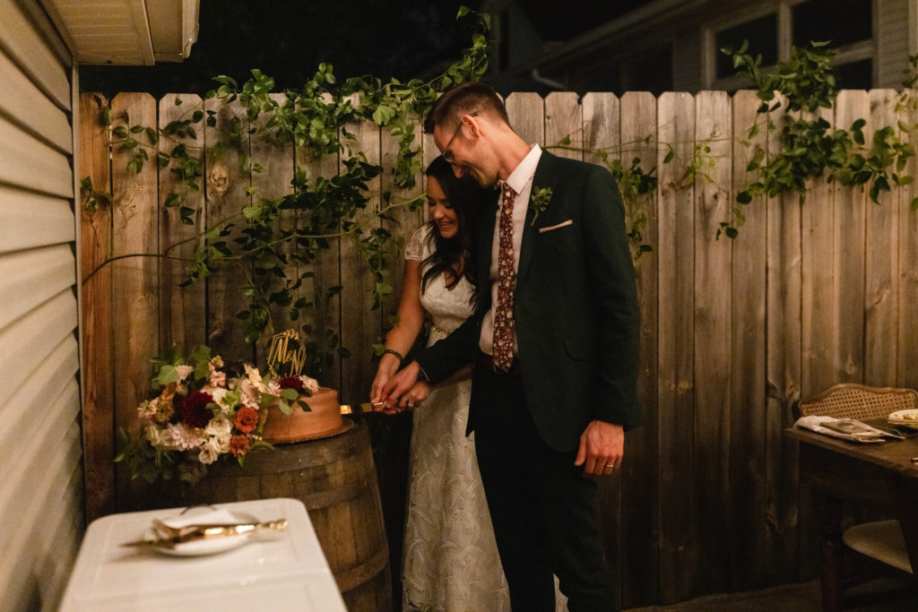 Bride and groom slicing their simple chocolate frosted cake adorned with majolica roses, a burgundy dahlia, and a geometric topper with calligraphy initials. It was displayed on a rustic barrel for this micro backyard wedding.